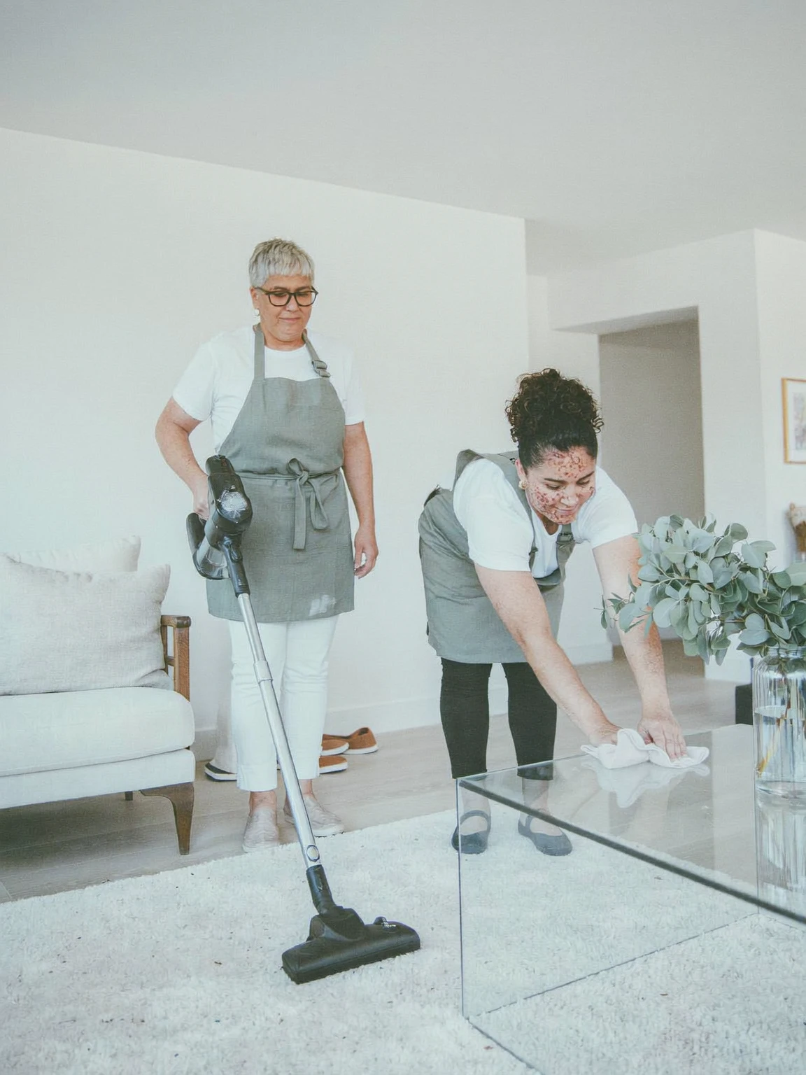 Two cleaners at work together, one vacuuming a cream rug and one wiping a glass coffee table
